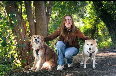 A smiling woman wearing glasses crouches between two medium-sized dogs on a woodland trail.