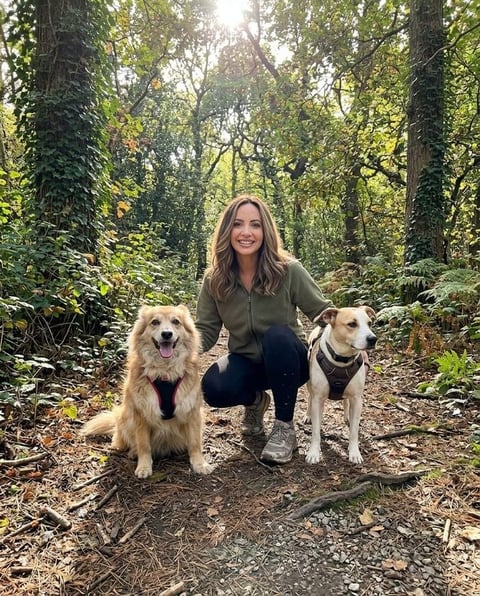 A woman crouches on a dirt path in a sun-dappled forest, smiling at the camera, flanked by two dogs wearing harnesses. The dog on the left is fluffy and light brown, while the dog on the right is white with brown patches.