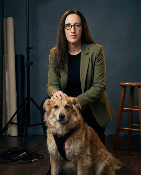 A woman with long dark hair and glasses, wearing a green blazer, kneels beside a medium-sized, fluffy brown dog wearing a black and red harness. The woman's hand rests on the dog's head. The background is a dark studio setting with photography equipment and a wooden stool visible.