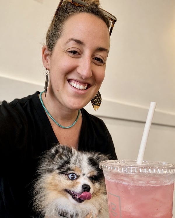 Eine lächelnde Frau posiert drinnen neben einem Blue-Merle-Zwergspitz mit Heterochromie und einem eisgekühlten pinken Getränk auf dem Tisch.