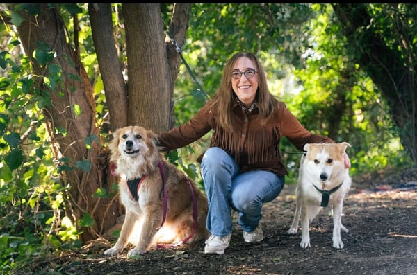 Eine lächelnde Frau mit Brille hockt zwischen zwei mittelgroßen Hunden auf einem Waldweg.